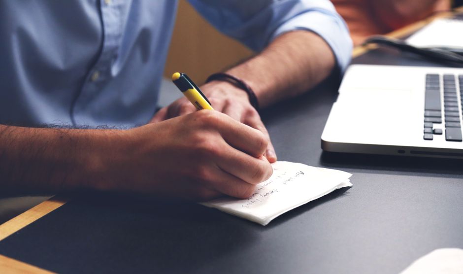 Close-up of a hand writing notes with a pen next to a laptop, representing original content creation