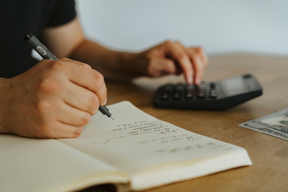 Close-up of hands writing calculations in a notebook next to a calculator